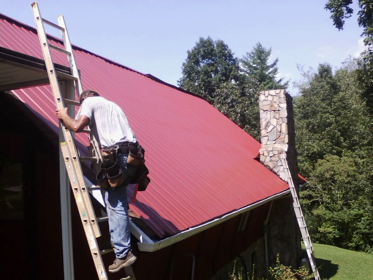 Roofer on a ladder beside a red metal roof and stone chimney — Soffit & Fascia Repair by Roofing Brothers in Parker City